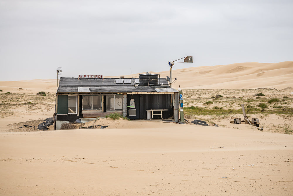 Filthy Phills Ranch at Tin City, sand drifts against the base of a low shack built from corrugated iron, timber cladding.