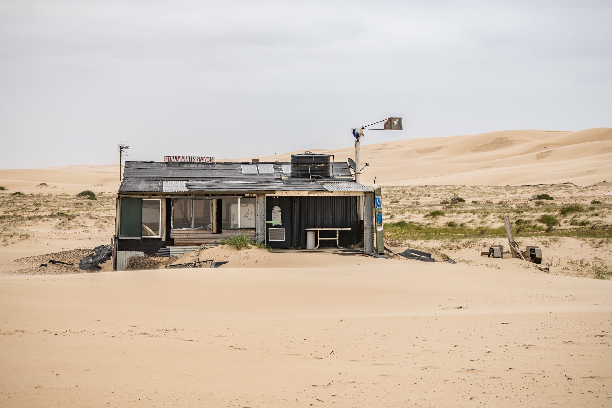 Filthy Phills Ranch at Tin City, sand drifts against the base of a low shack built from corrugated iron, timber cladding.