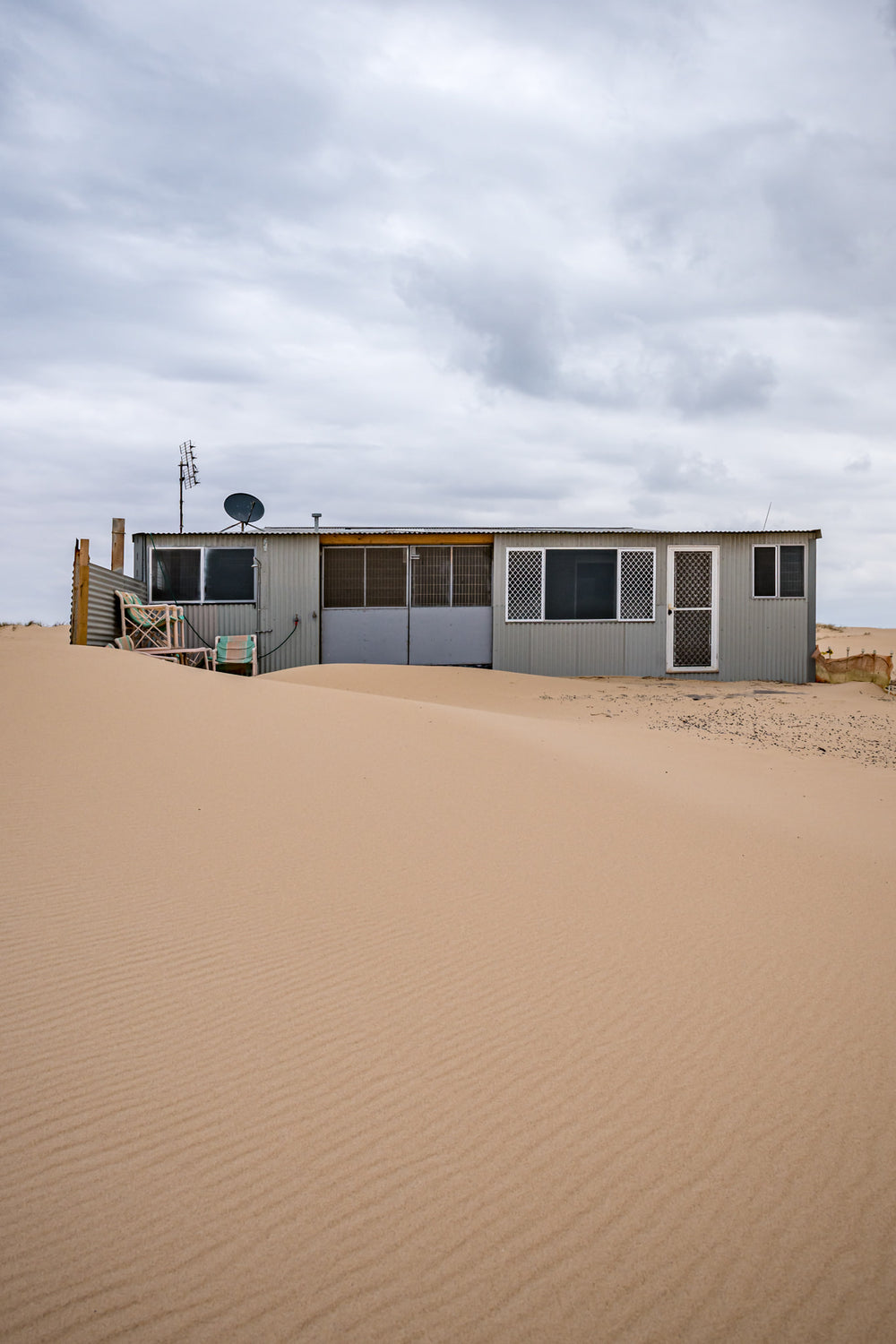 Ripples at Tin City, sand rises in a smooth, wind-rippled drift against a low corrugated iron shack.