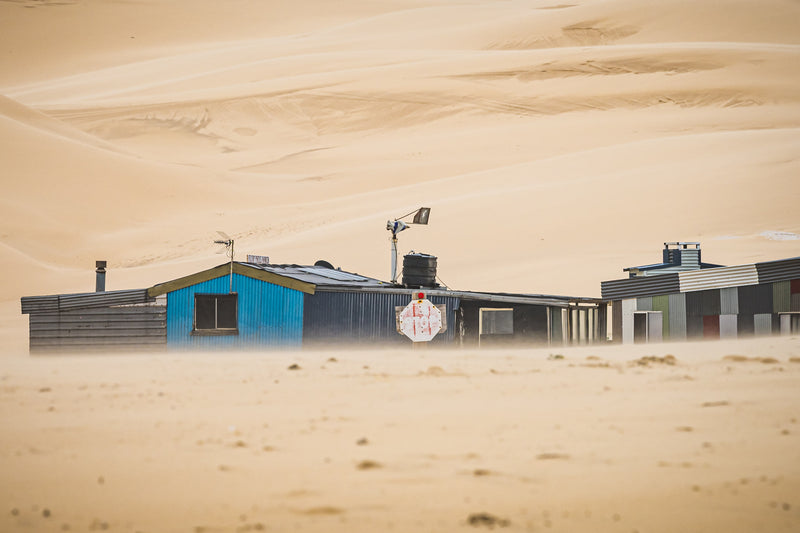 Sandstorm at Tin City, sand drives low across the ground, blurring the base of a row of shacks at Tin City.