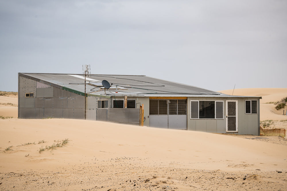 Sloped Roof at Tin City, a corrugated iron shack sits half-swallowed by sand.