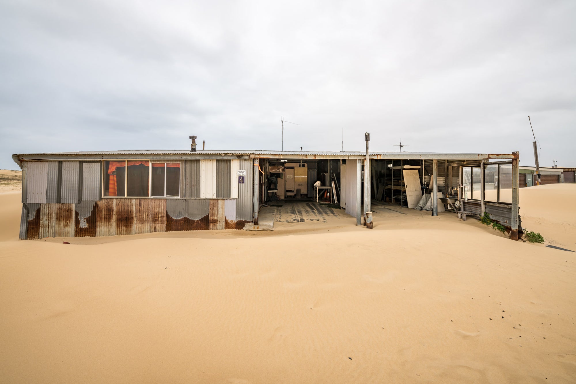 The Garage at Tin City, sand presses against the lower walls of a long, low shack at Tin City.