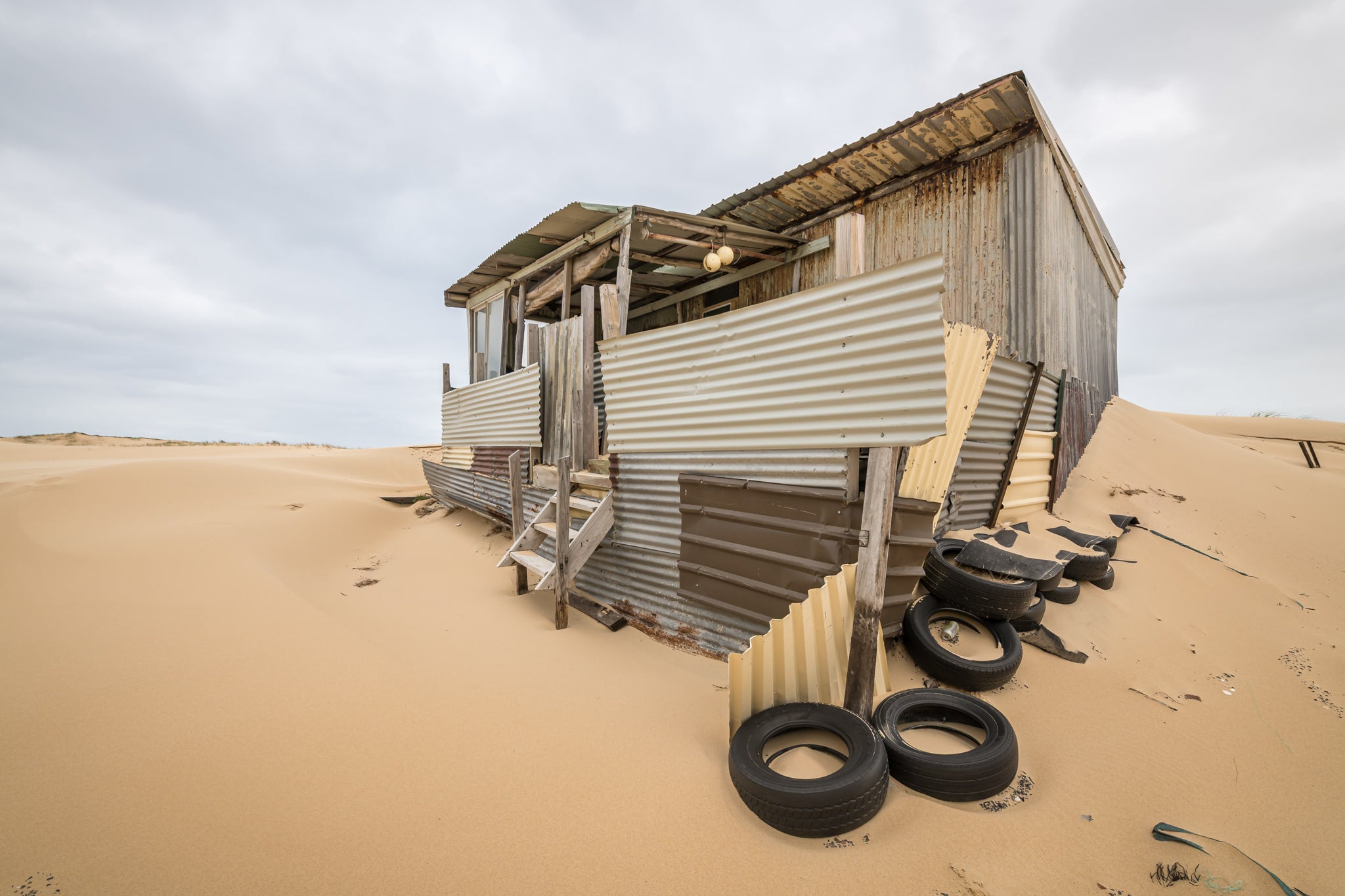 Tyres at Tin City, a corrugated iron shack lists sideways into a dune, its verandah posts leaning at odd angles.