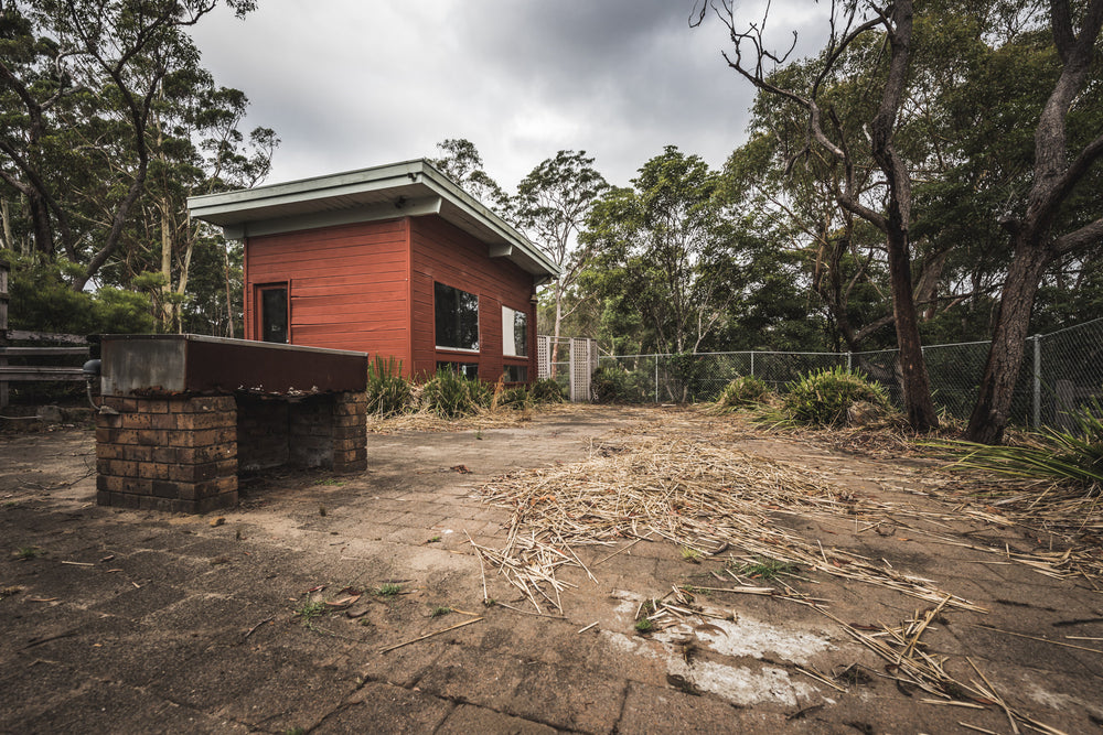Bbq Area at Waratah Park, a red-painted timber cabin sits on a wide concrete paver courtyard surrounded by eucalyptus bush.