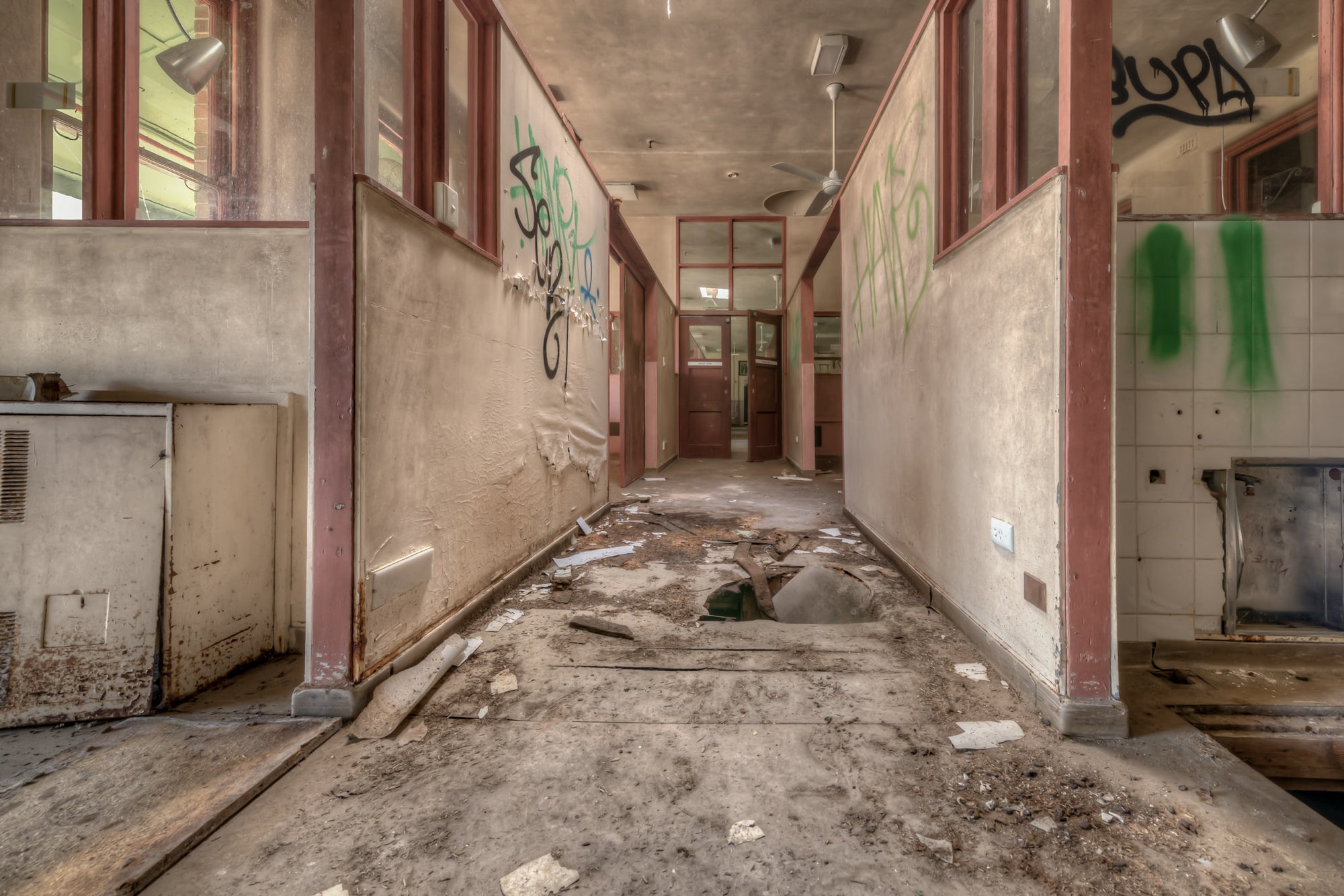 Fallen Through at Waterfall Sanatorium, a corridor runs between partitioned rooms, their maroon-trimmed frames holding.