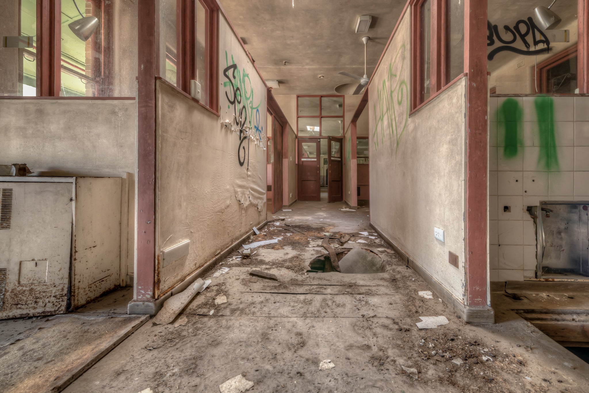 Fallen Through at Waterfall Sanatorium, a corridor runs between partitioned rooms, their maroon-trimmed frames holding.