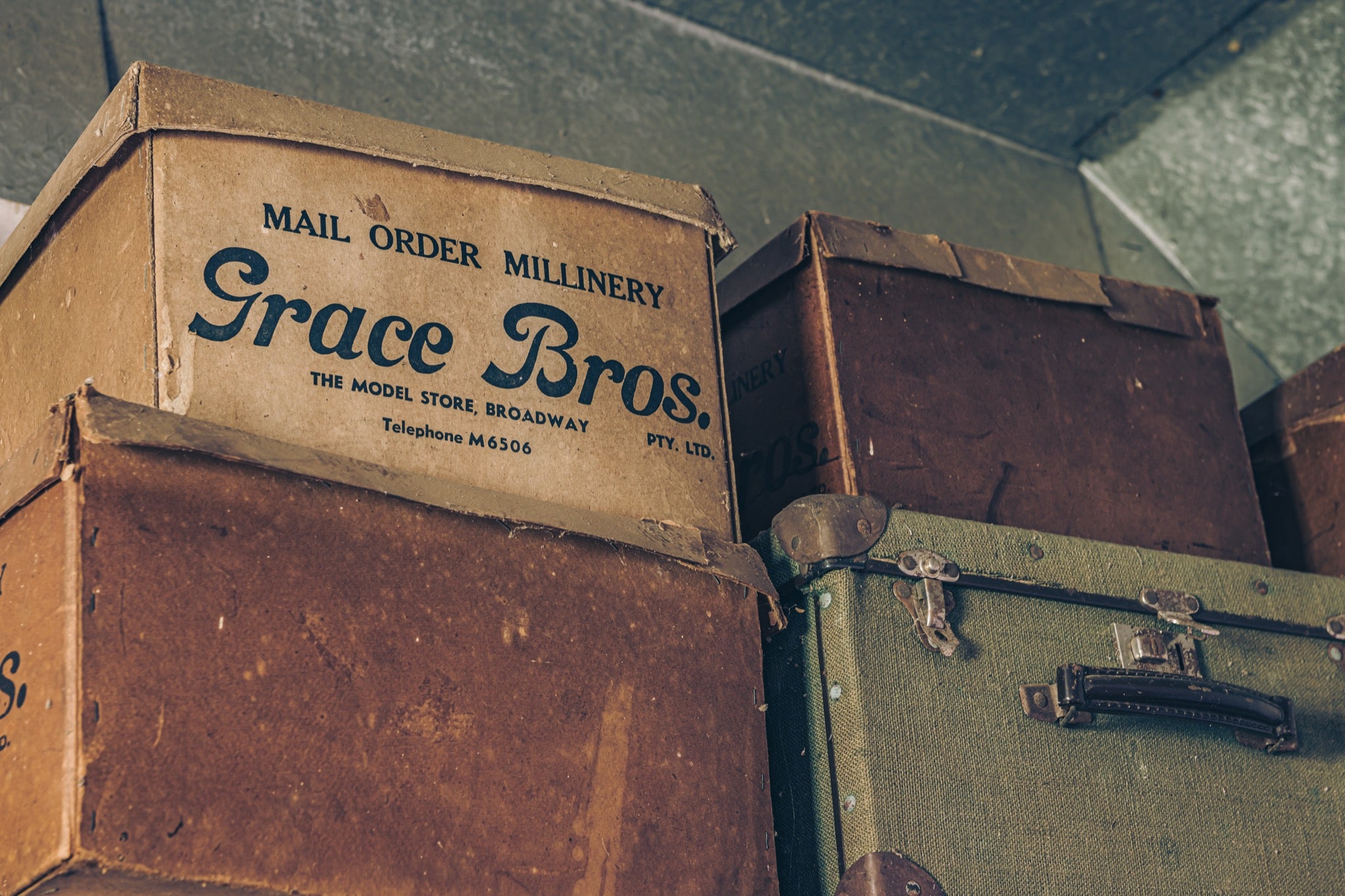 Grace Bros. hatboxes stacked beside a green canvas suitcase with metal latches and a leather handle, photographed at Moruya.