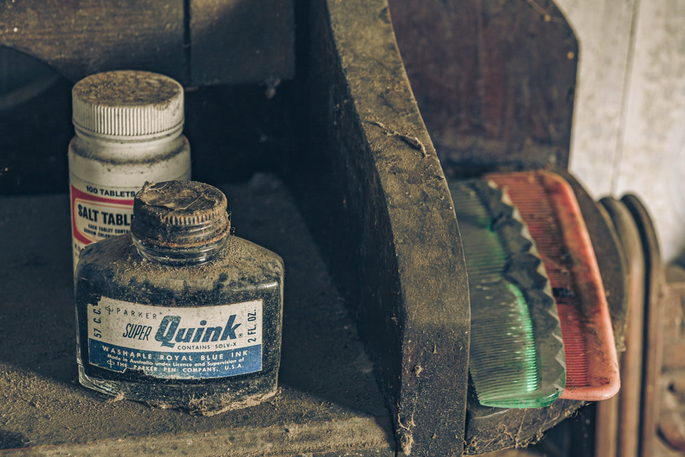 A dust-covered glass ink bottle and white tablet jar on a grimy timber shelf at Woolla, with a green and pink comb resting beside them.