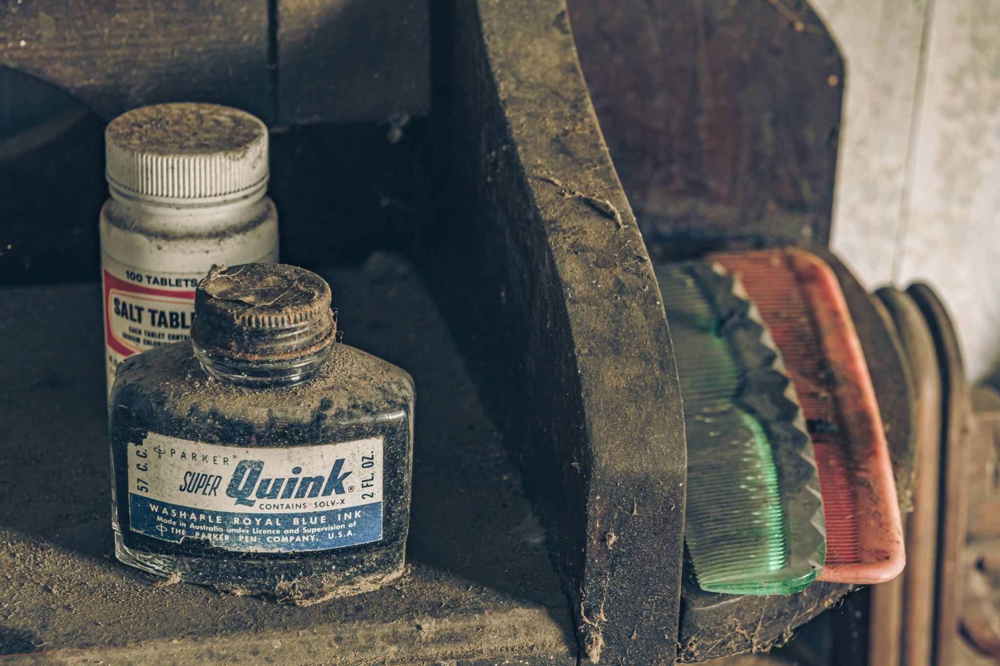 A dust-covered glass ink bottle and white tablet jar on a grimy timber shelf at Woolla, with a green and pink comb resting beside them.