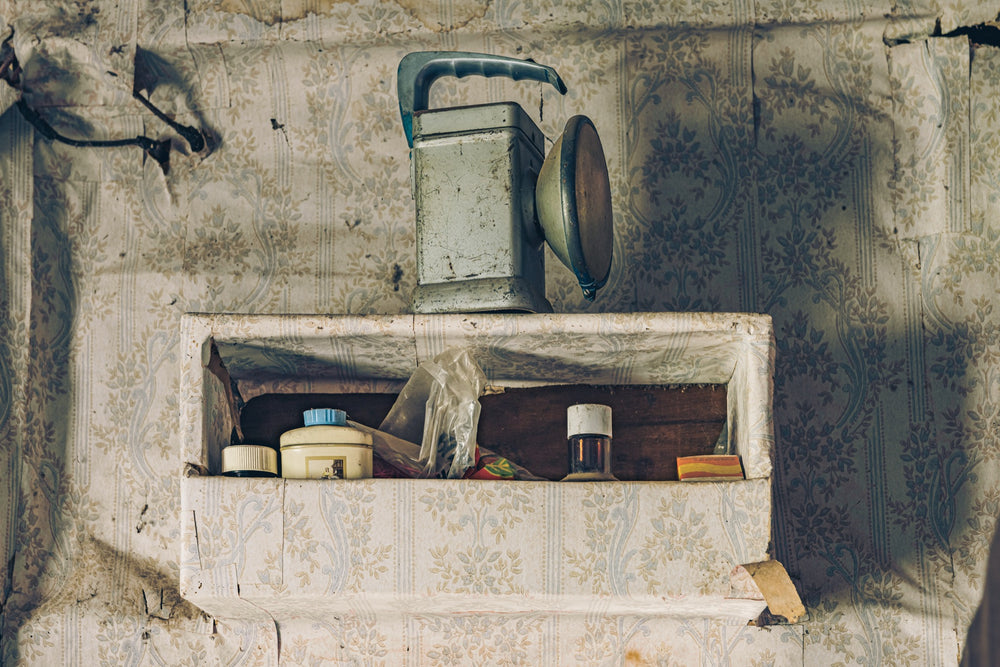 A metal lantern with its reflector open sits on a recessed shelf set into torn floral wallpaper at Woolla, with small jars, a lidded container, a plastic bag, and a matchbox arranged below it.