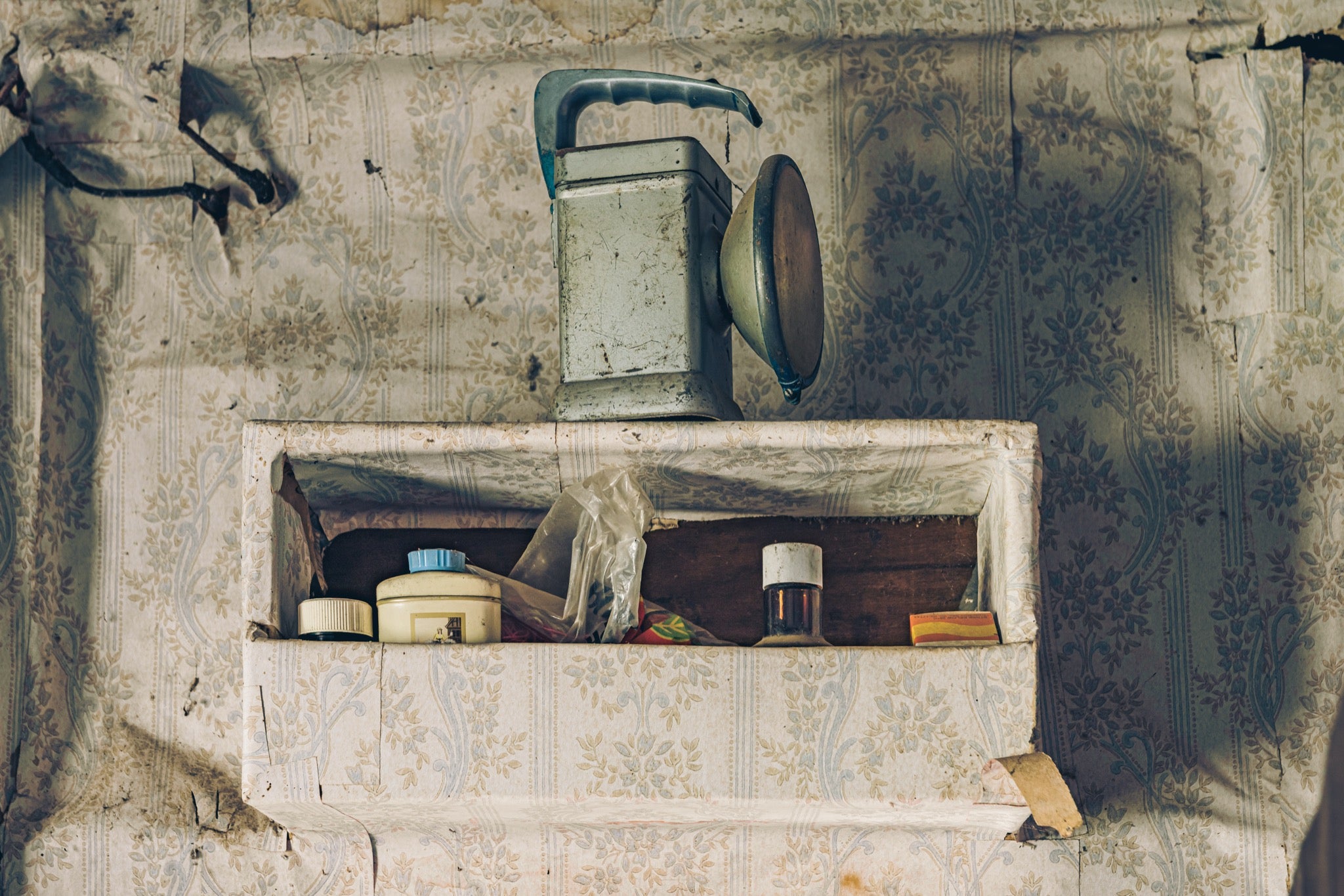 A metal lantern with its reflector open sits on a recessed shelf set into torn floral wallpaper at Woolla, with small jars, a lidded container, a plastic bag, and a matchbox arranged below it.
