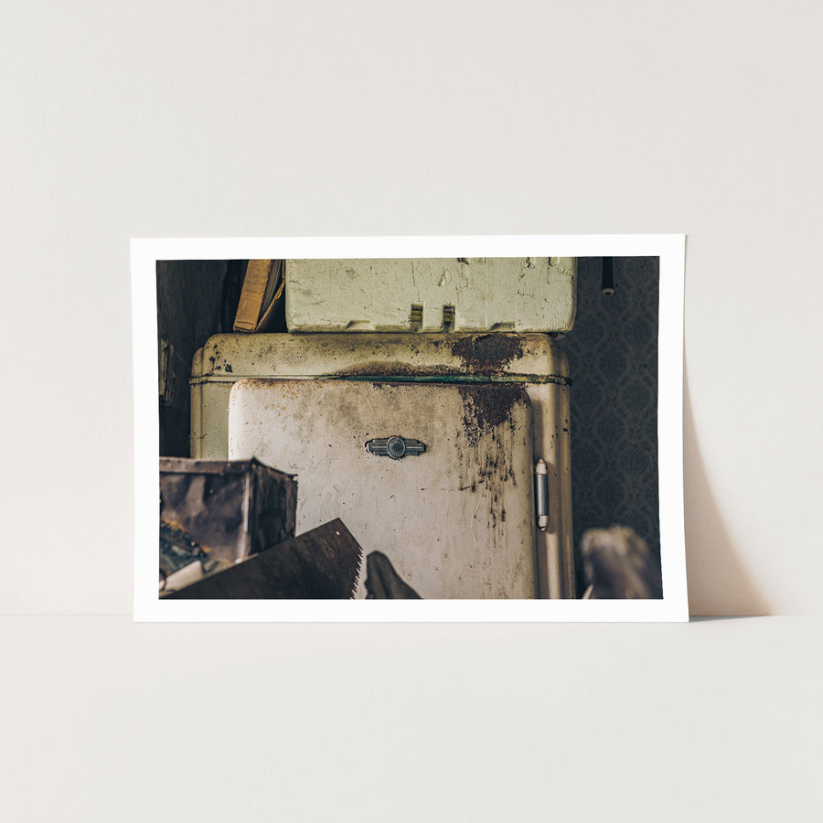 An old enamel refrigerator with a rusted top edge and chrome latch stands against patterned wallpaper at Woolla in the Deua River Valley, a handsaw and cardboard boxes on the floor beside it.