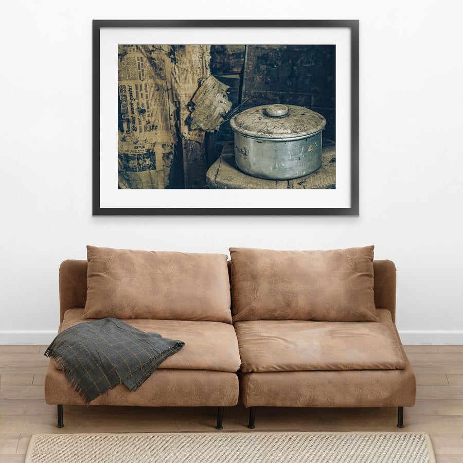 A dust-covered aluminium saucepan and domed lid on a worn wooden surface inside a slab hut at Woolla in the Deua River Valley, with yellowed newspaper lining the timber walls behind.