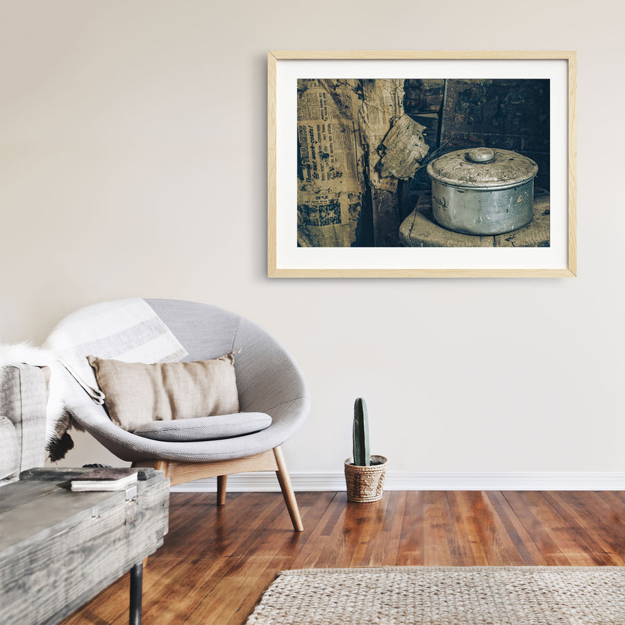 A dust-covered aluminium saucepan and domed lid on a worn wooden surface inside a slab hut at Woolla in the Deua River Valley, with yellowed newspaper lining the timber walls behind.