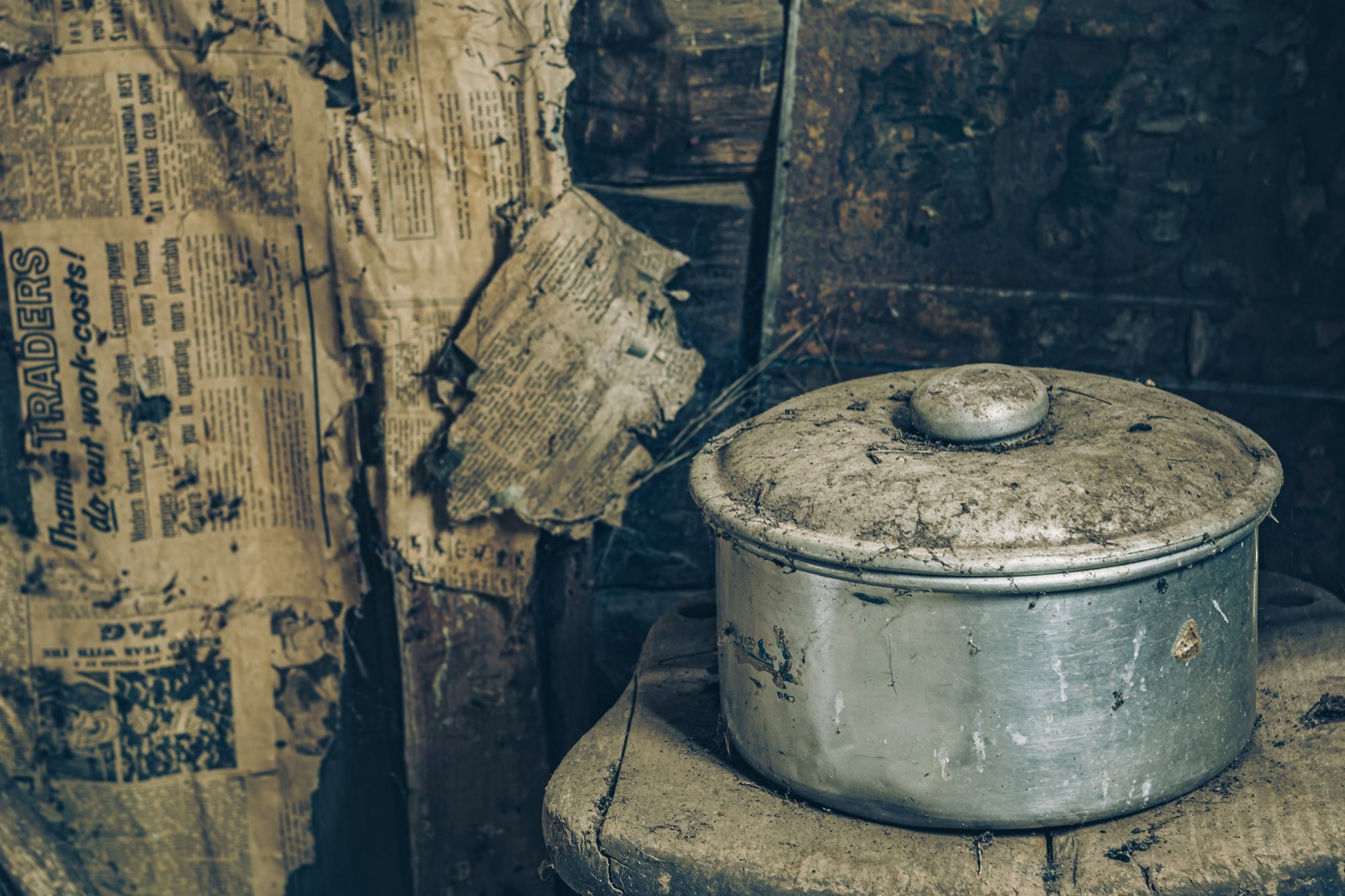 A dust-covered aluminium saucepan and domed lid on a worn wooden surface inside a slab hut at Woolla in the Deua River Valley, with yellowed newspaper lining the timber walls behind.