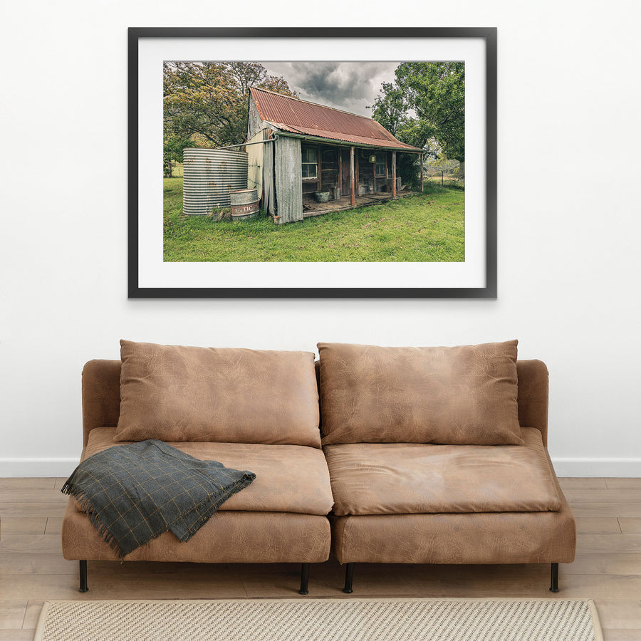 The weathered timber slab hut and covered front verandah at Woolla, with a galvanised corrugated water tank and steel drum standing at the gable end on mown grass among trees near Braidwood.