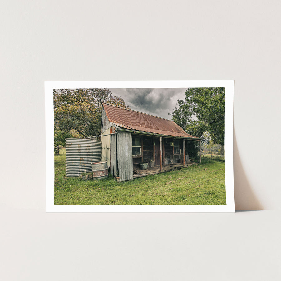 The weathered timber slab hut and covered front verandah at Woolla, with a galvanised corrugated water tank and steel drum standing at the gable end on mown grass among trees near Braidwood.