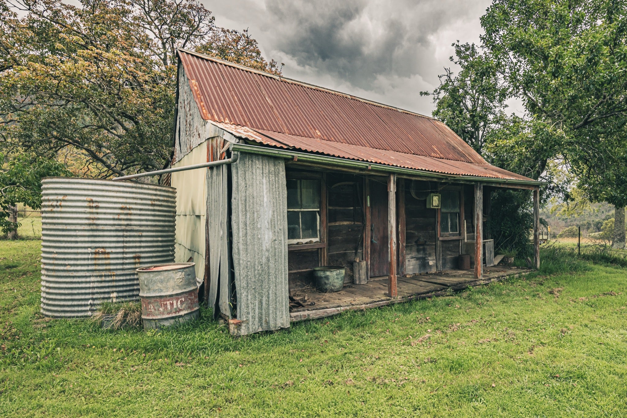 The weathered timber slab hut and covered front verandah at Woolla, with a galvanised corrugated water tank and steel drum standing at the gable end on mown grass among trees near Braidwood.
