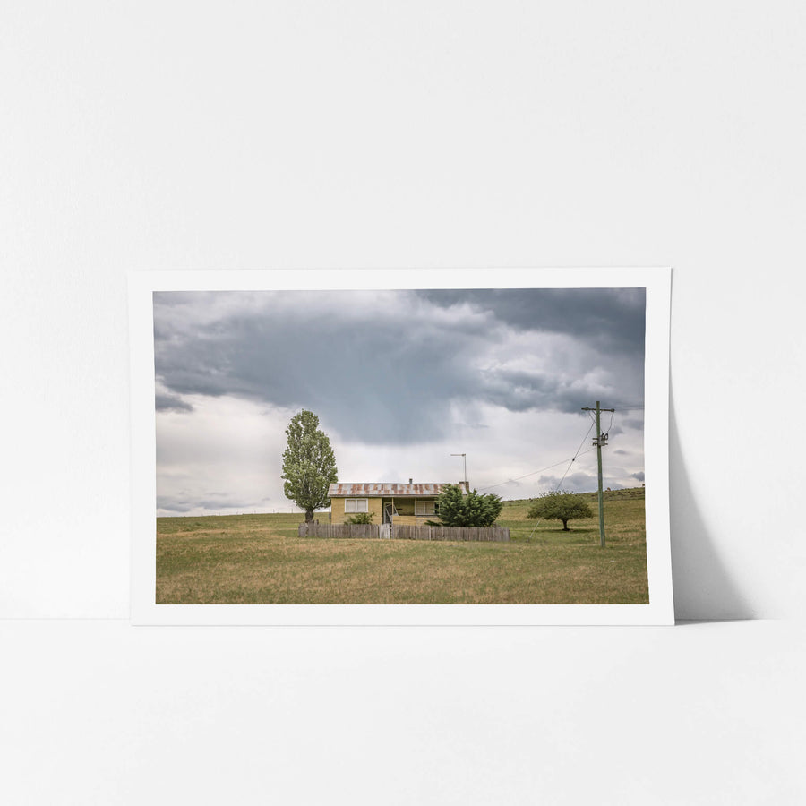 Home On the Range at A Place to Call Home, a long-vacant, lone farm cottage sits in a field beneath a distant rain cloud.