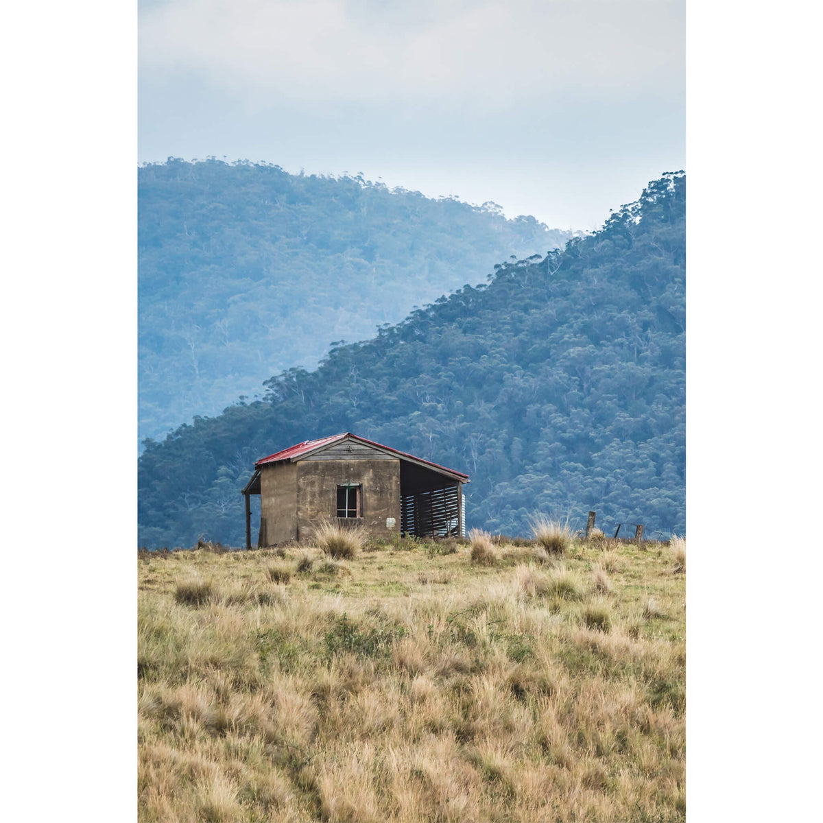 A solitary weathered house with a red roof perched on a hill, surrounded by high country grasslands and mountains in the background.