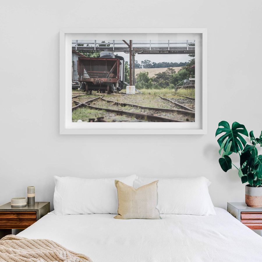 Upline at Bombala Station, a rusted hopper wagon sits low on the sidings beneath a steel and timber footbridge.