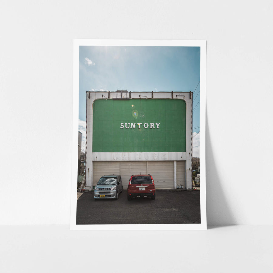Suntory at Streetscapes of Yubari, a broad green façade rises two storeys above a small asphalt lot.