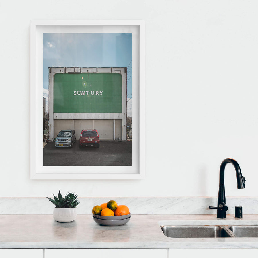 Suntory at Streetscapes of Yubari, a broad green façade rises two storeys above a small asphalt lot.