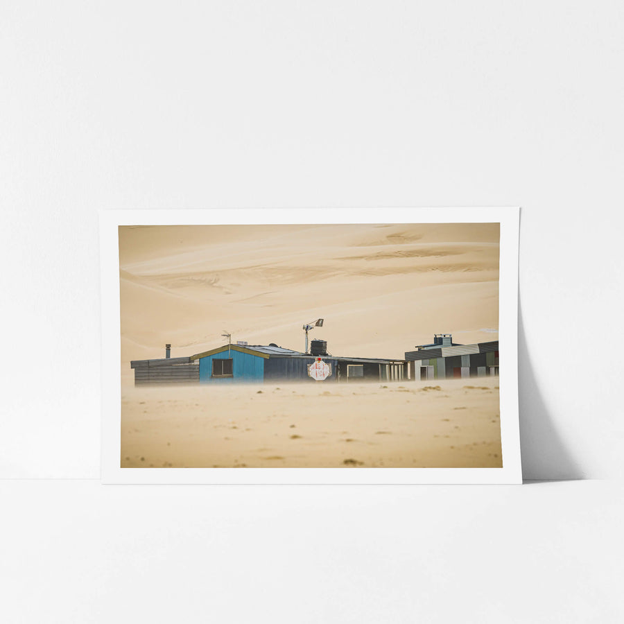 Sandstorm at Tin City, sand drives low across the ground, blurring the base of a row of shacks at Tin City.